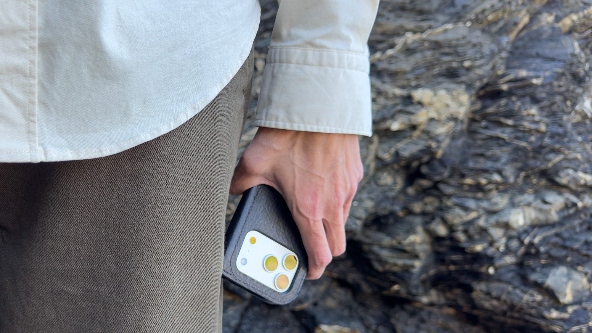 Lifestyle — iPhone 17 Series in Classic 5.0 leather folio case wallet, in-hand carry by rocky backdrop; full-grain Italian leather, MagSafe-ready minimal profile