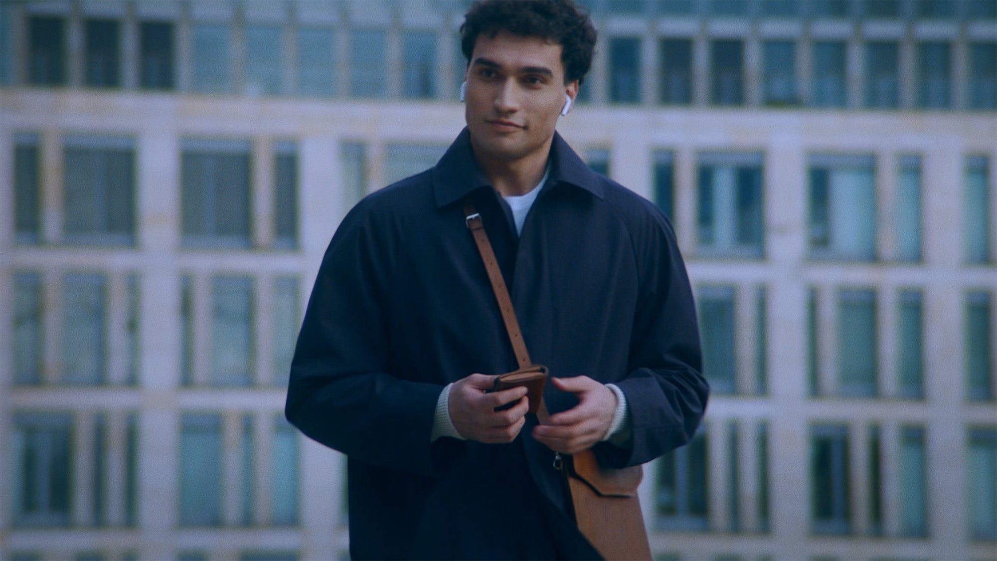 Man holding a brown leather bifold AirTag wallet, standing outdoors in front of a modern building. The AirTag wallet combines style with tracking functionality