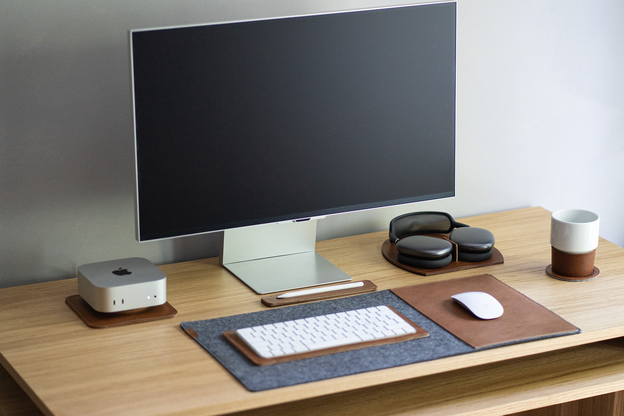 AirPods Max on a brown leather rest station beside a monitor; felt desk pad with leather mouse area on a wooden desk.