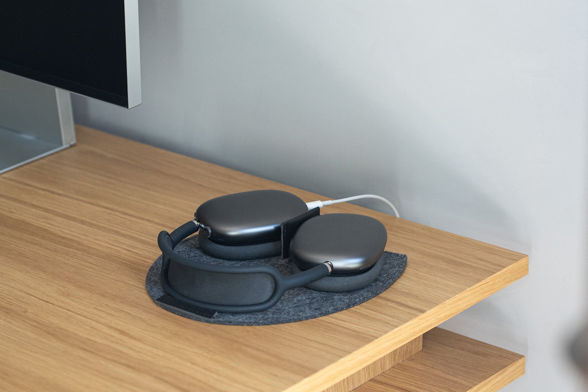AirPods Max on grey wool felt rest station with leather magnetic tab and cable pass-through; charging on a wooden desk.
