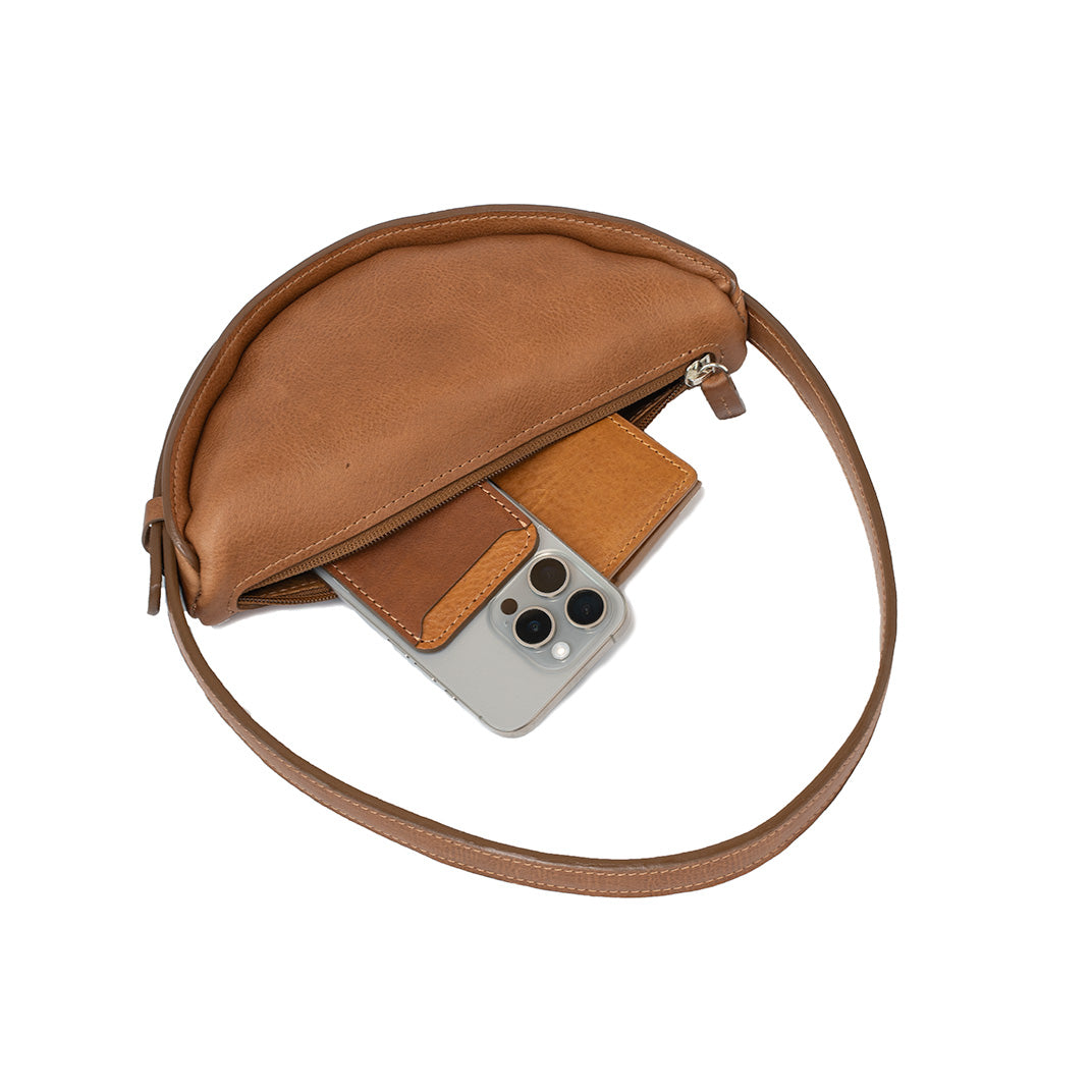 Brown leather half-moon bag opened, revealing spacious interior with essentials like a phone and wallet inside