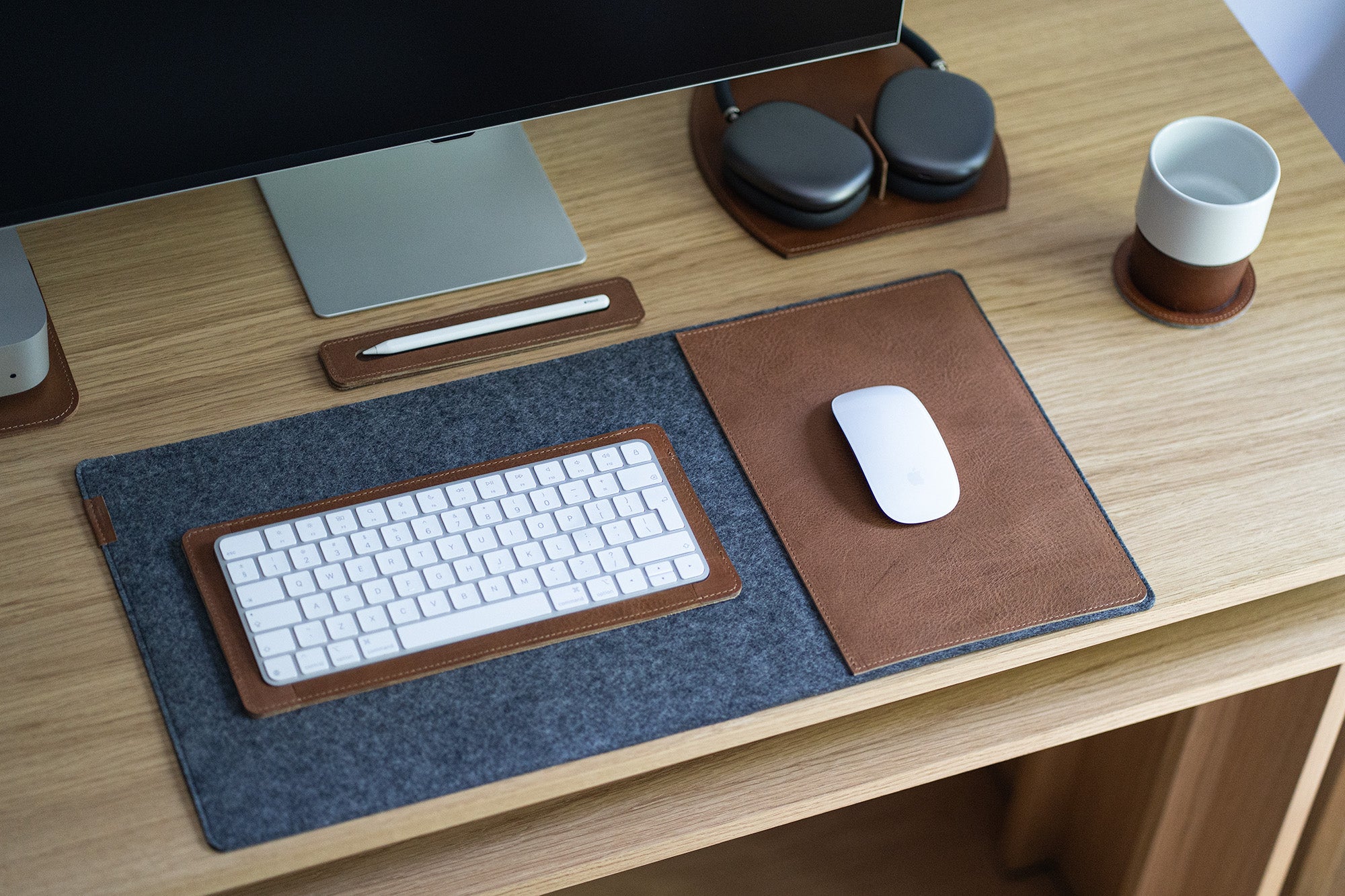 Grey felt desk pad with brown leather mouse insert in a minimalist setup; Magic Keyboard, Magic Mouse, and Apple Pencil tray.
