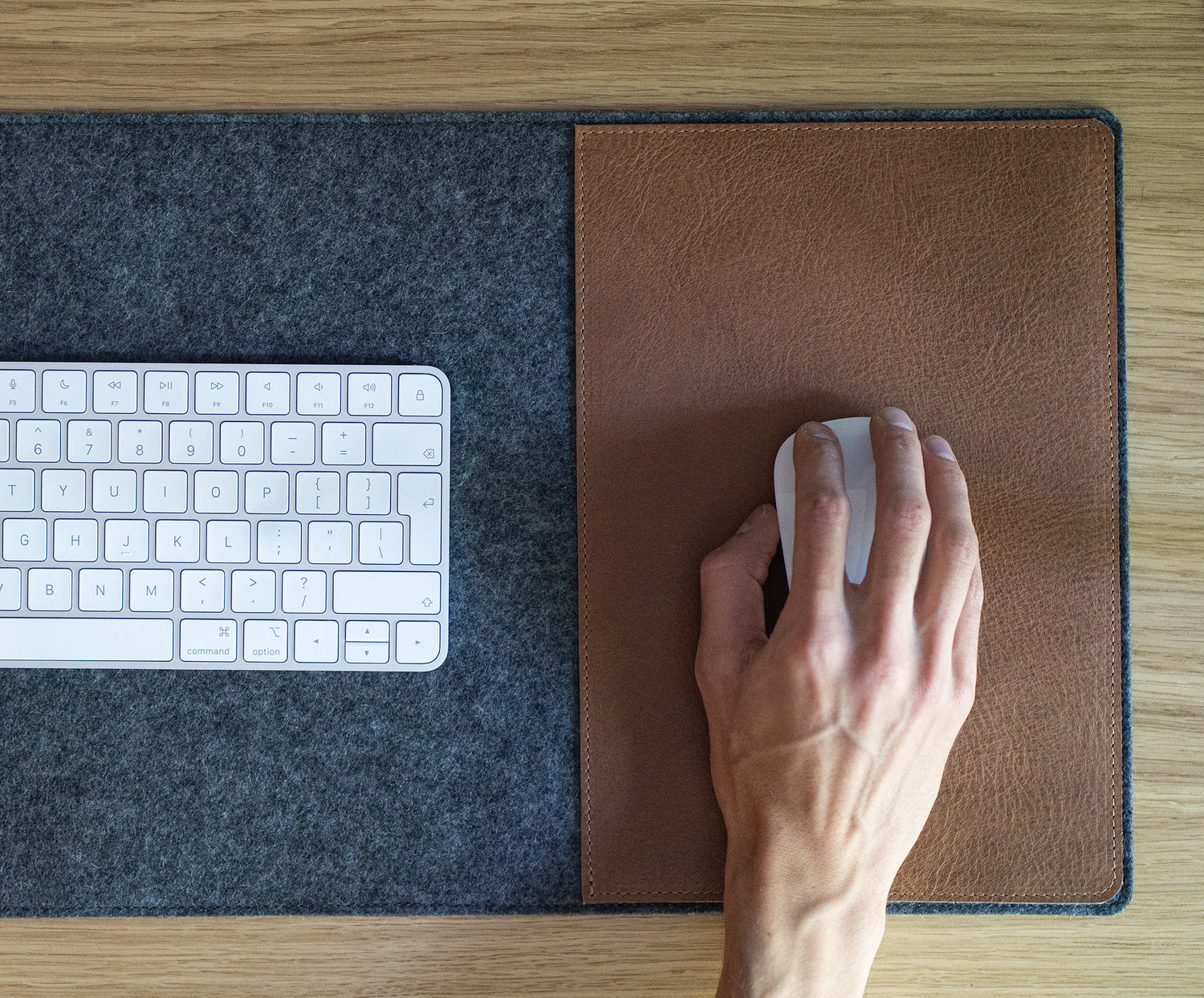 Hand using a white Magic Mouse on the brown leather mouse area of a grey felt desk pad; keyboard on the felt base.