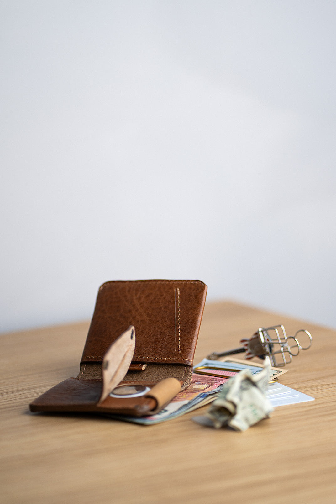 Minimalist leather AirTag wallet with cash strap, holding US dollars and euro banknotes, placed on a wooden desk surface