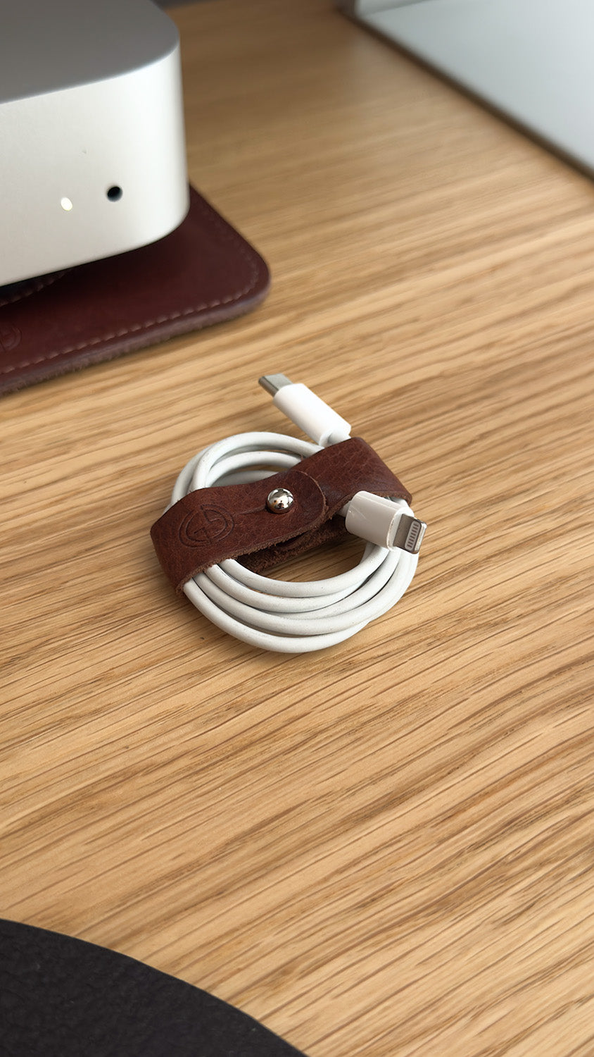 Close-up of leather cable organizer securing white USB-C to Lightning cable on wooden desk next to Mac mini