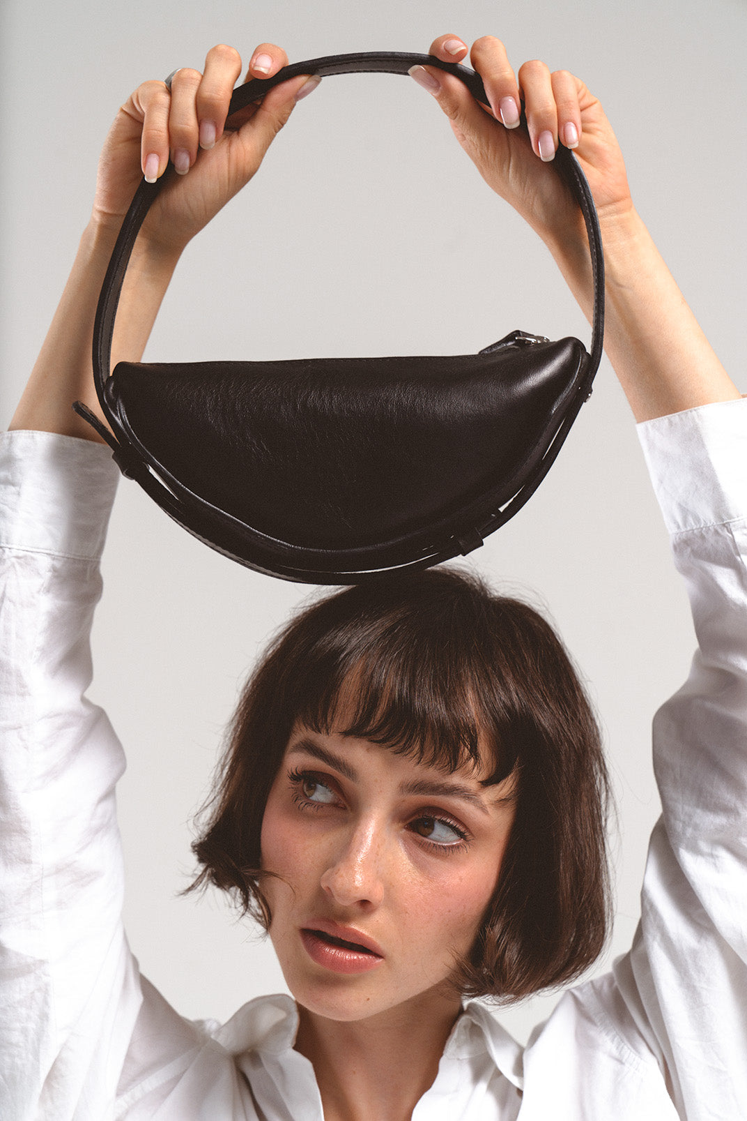 Leather shoulder bag Thales in black, held overhead by a model, showcasing its crescent shape and smooth leather finish