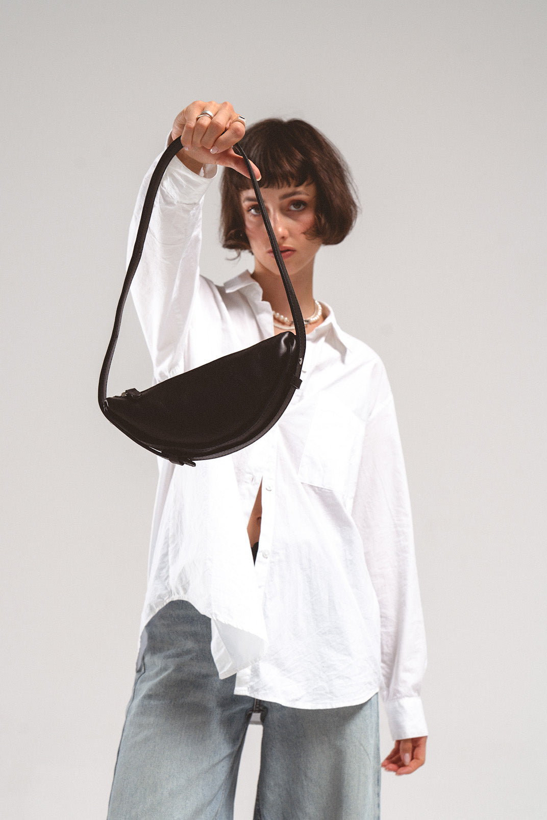 Leather shoulder bag Thales in black, held forward by a model wearing a white shirt and jeans, highlighting its crescent shape and sleek desig