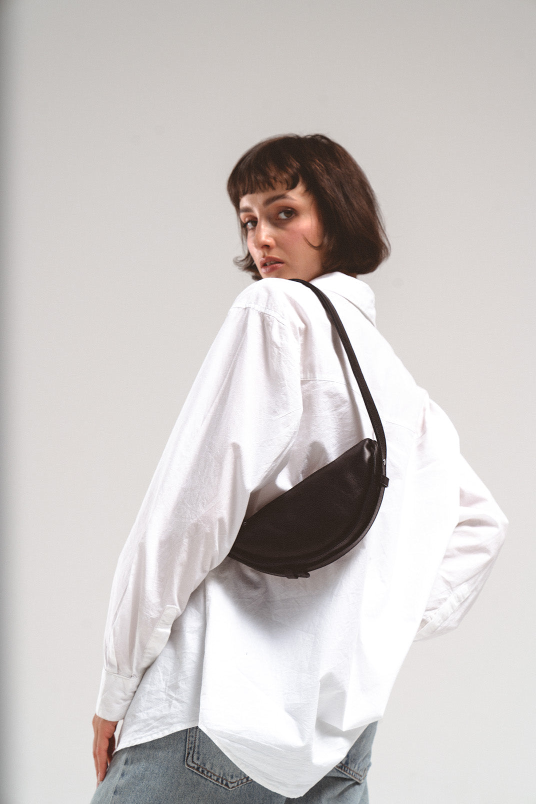 Leather shoulder bag Thales in black, worn over the shoulder by a model in a white shirt, showcasing the crescent shape and sleek design