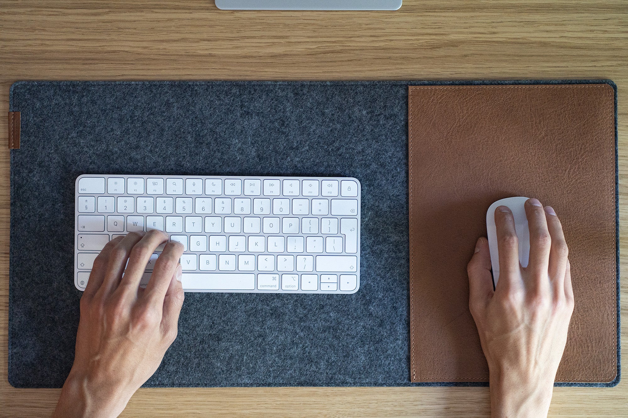 Grey wool felt desk pad with brown leather mouse insert; hands on keyboard and mouse on a wooden desk.