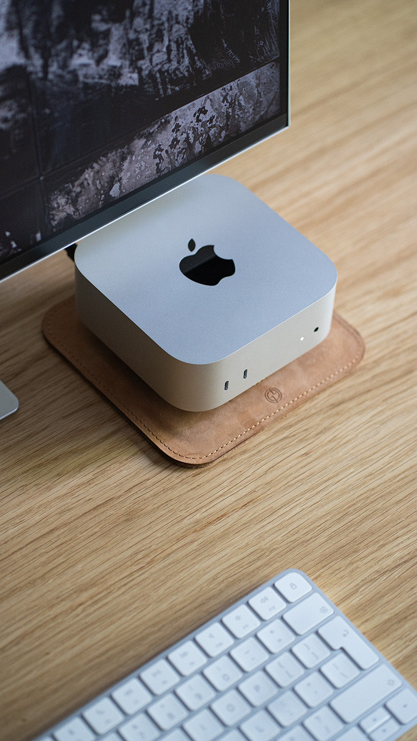 Top angled view of Mac Mini on The Pedestal leather stand, placed under monitor on wooden desk with Apple keyboard