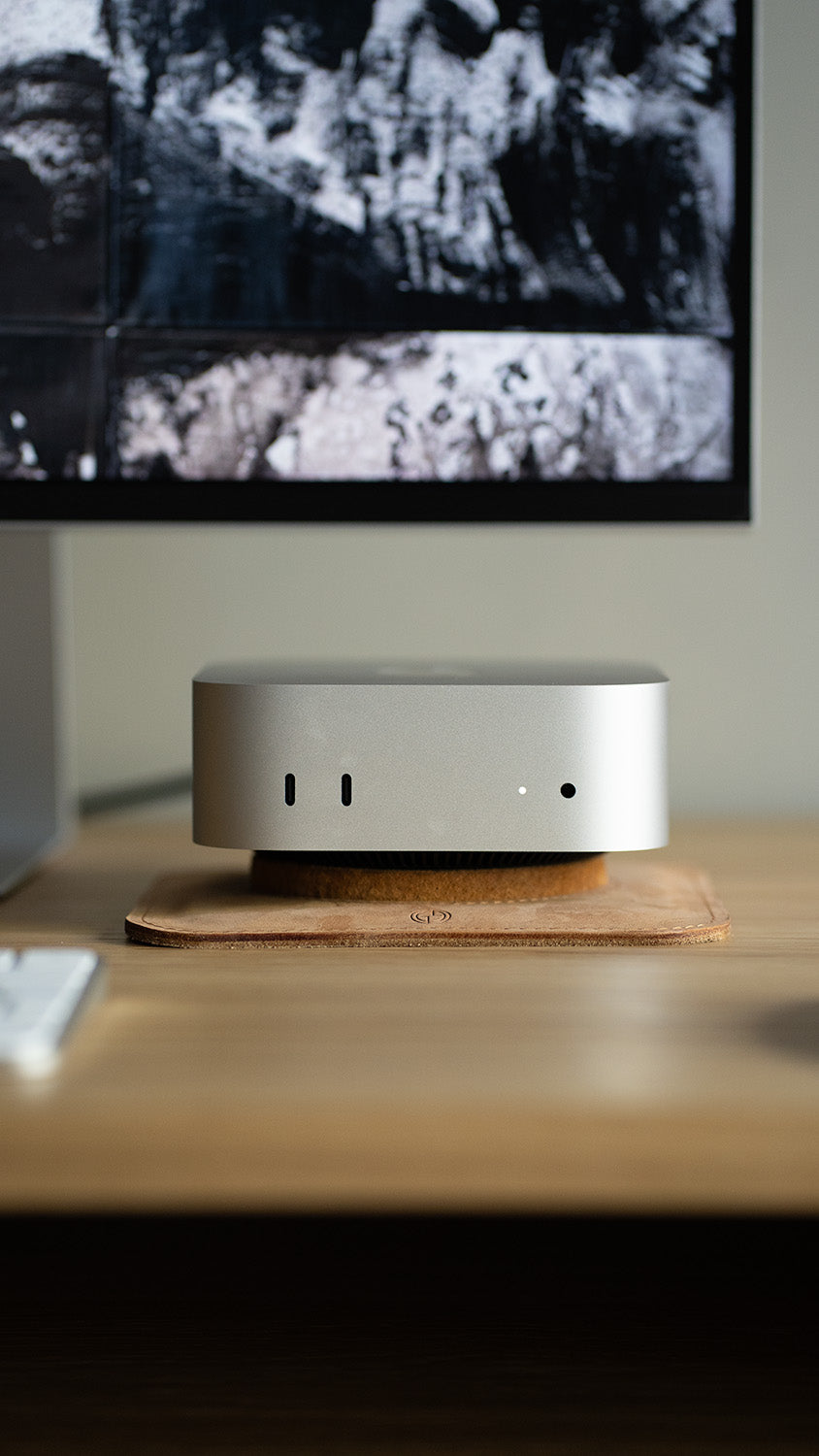 Close-up front view of Mac Mini on handcrafted leather pedestal stand, shown under monitor on wooden desk