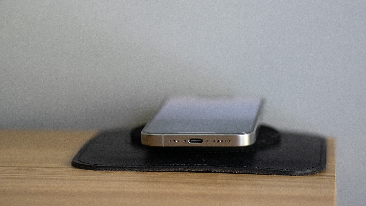 Minimalist MagSafe leather pad with iPhone resting – black charging mat shown from side on wooden workspace