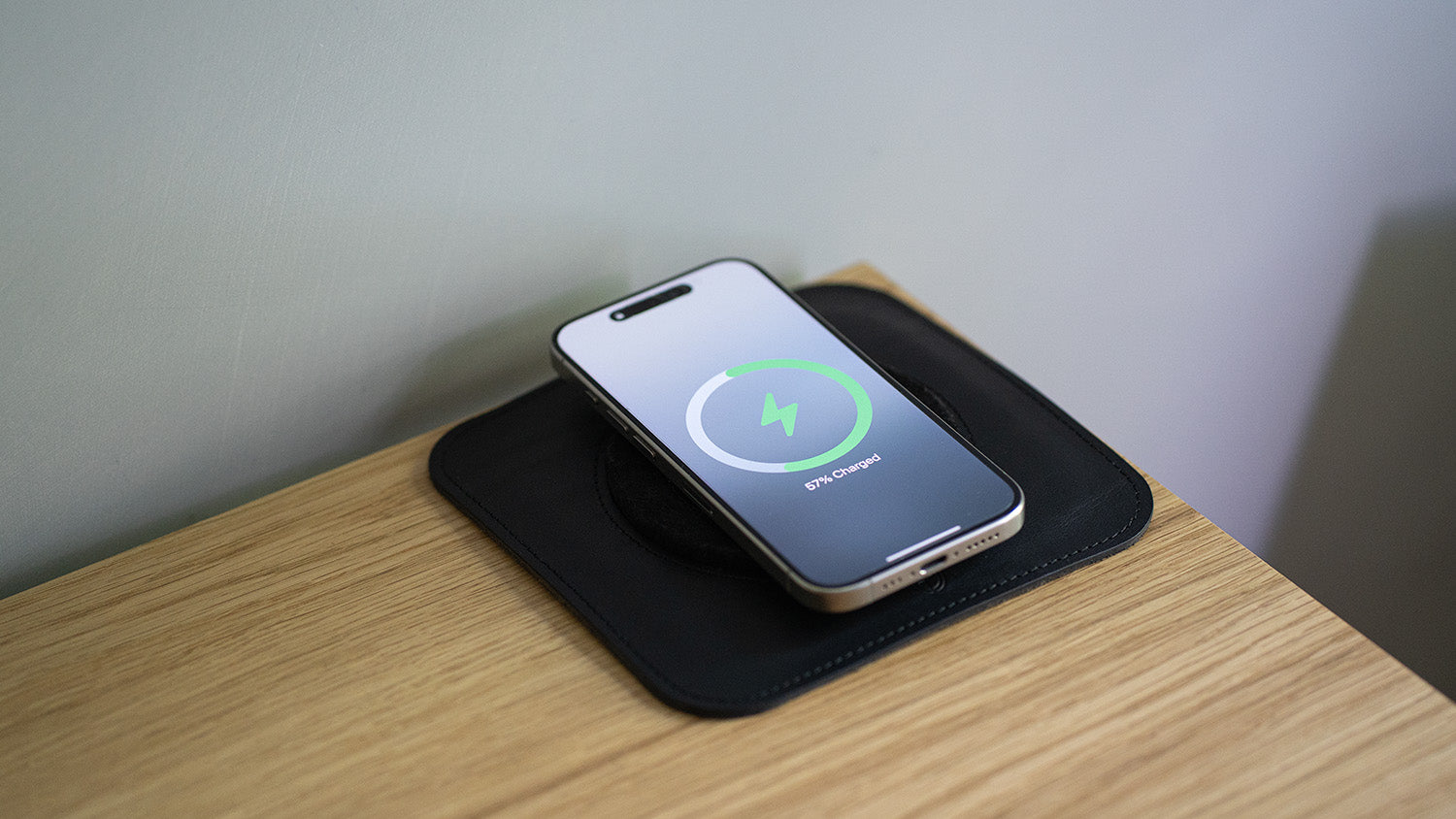 iPhone charging on black leather MagSafe rest mat placed on wooden desk, showing 57% charged status