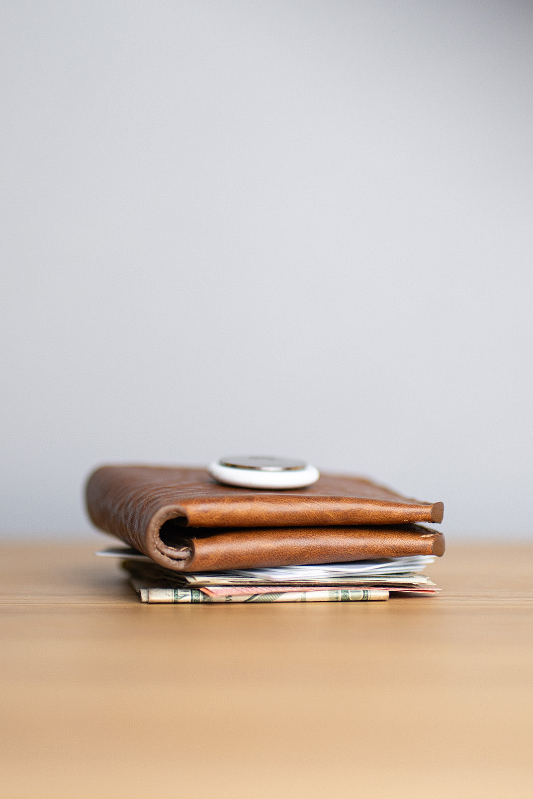 Slim leather AirTag billfold wallet with cash and cards, viewed from the side on a wooden desk