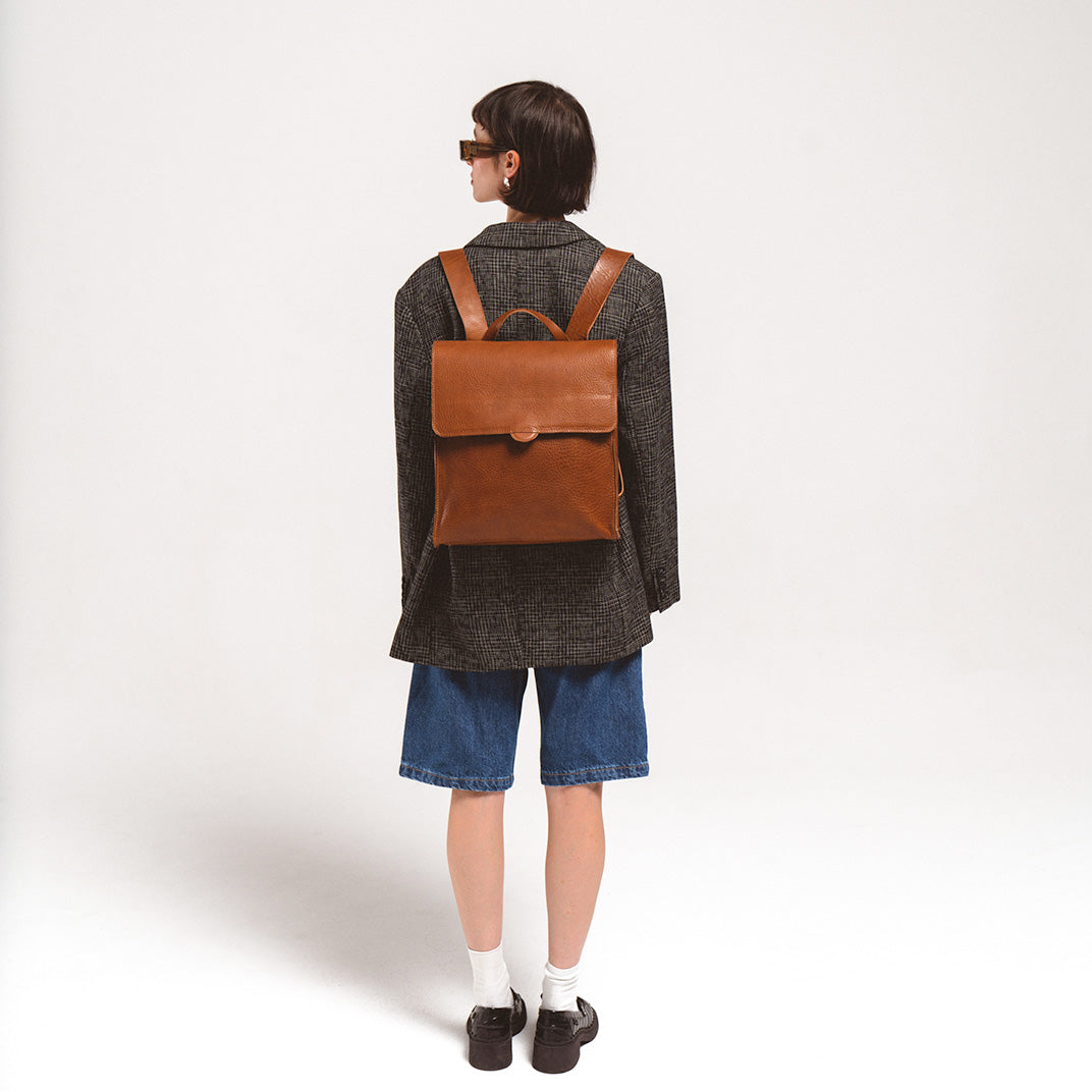 Leather backpack for woman elegantly carried by a young woman in a blazer and denim shorts, focusing on the smooth brown leather finish